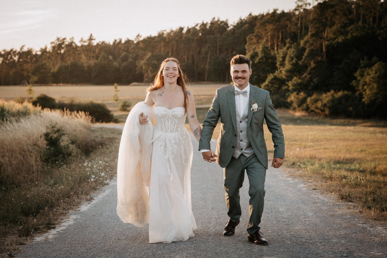 Brautpaar läuft im Abendlicht Hand in Hand einen Feldweg entlang