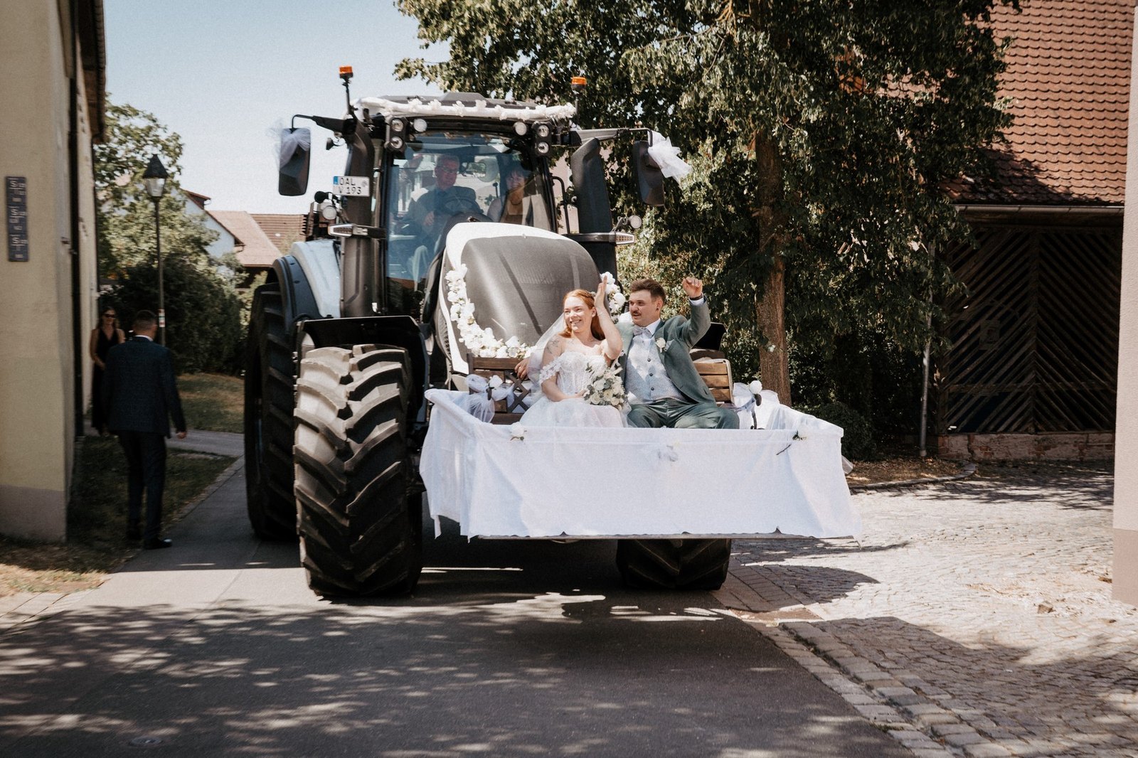 Brautpaar faehrt in der geschmueckten Frontschaufel eines grossen Traktors durch den Ort und winkt