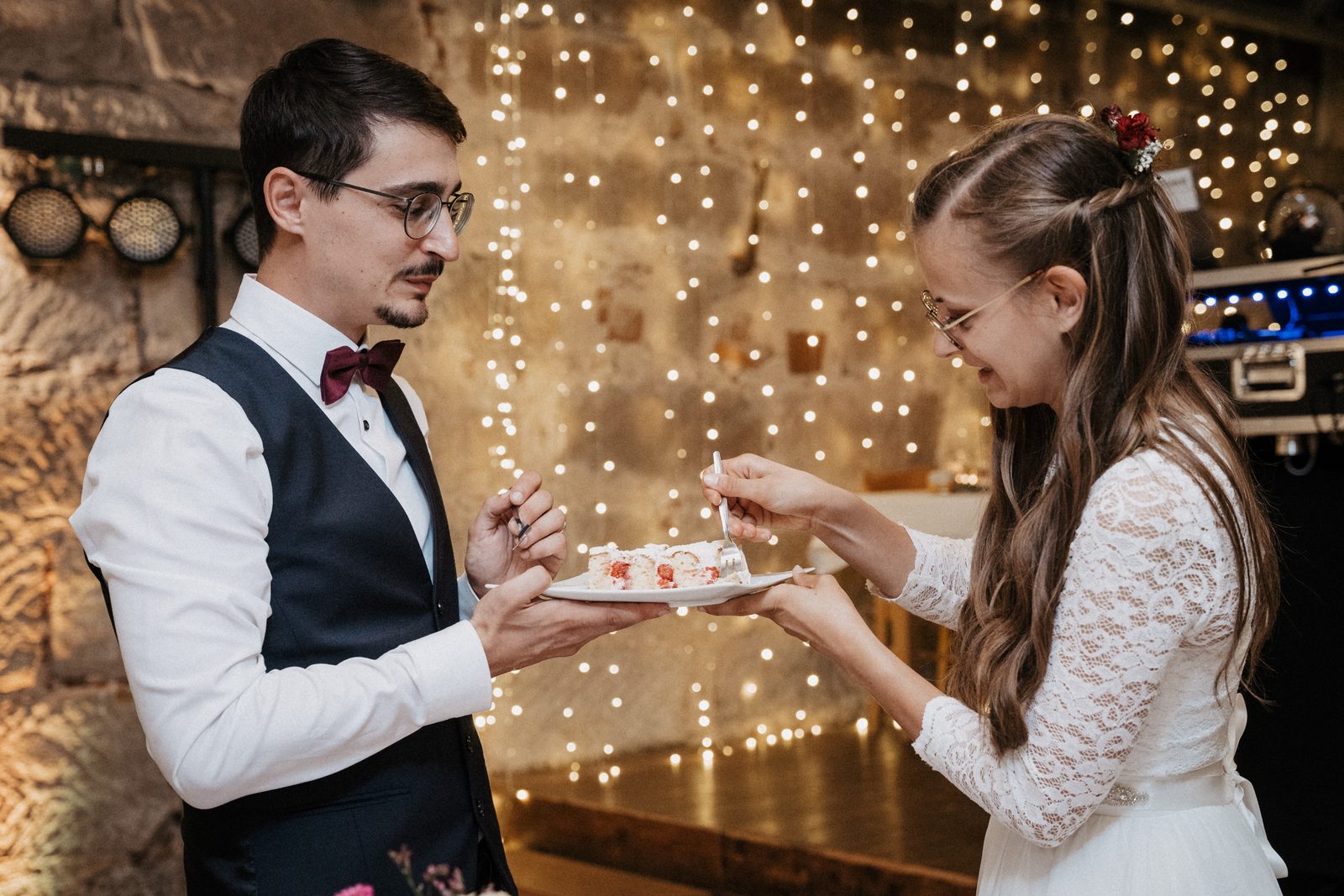Brautpaar teilt sich ein Stück Hochzeitstorte vor einer Wand aus Lichterketten