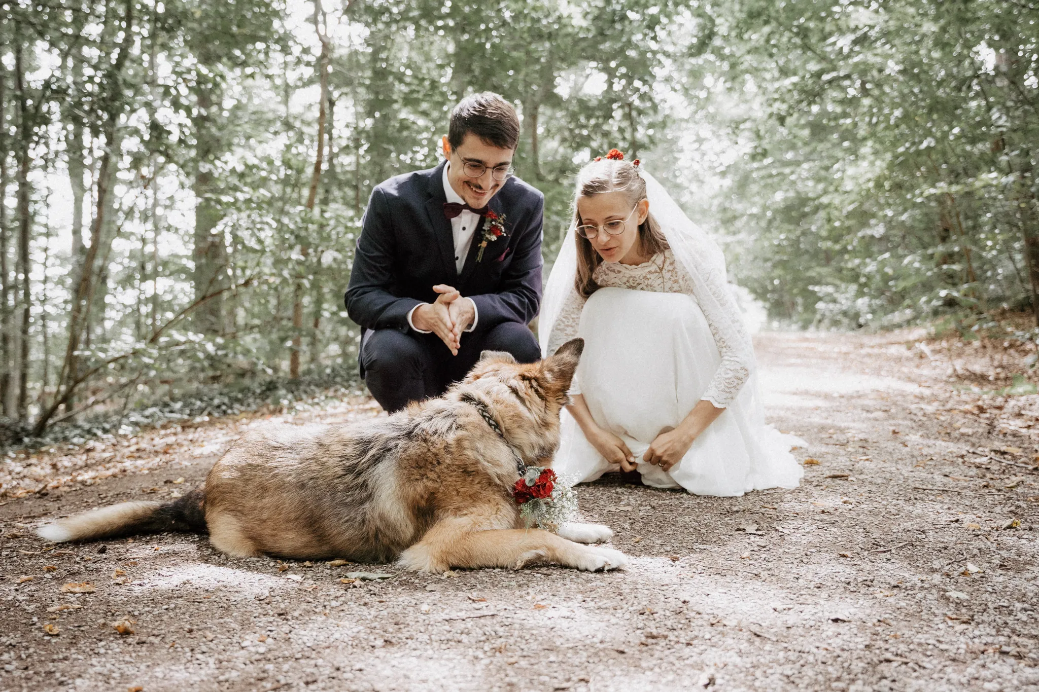 Brautpaar kniet im Wald neben seinem Hund, der einen Blumenkranz trägt