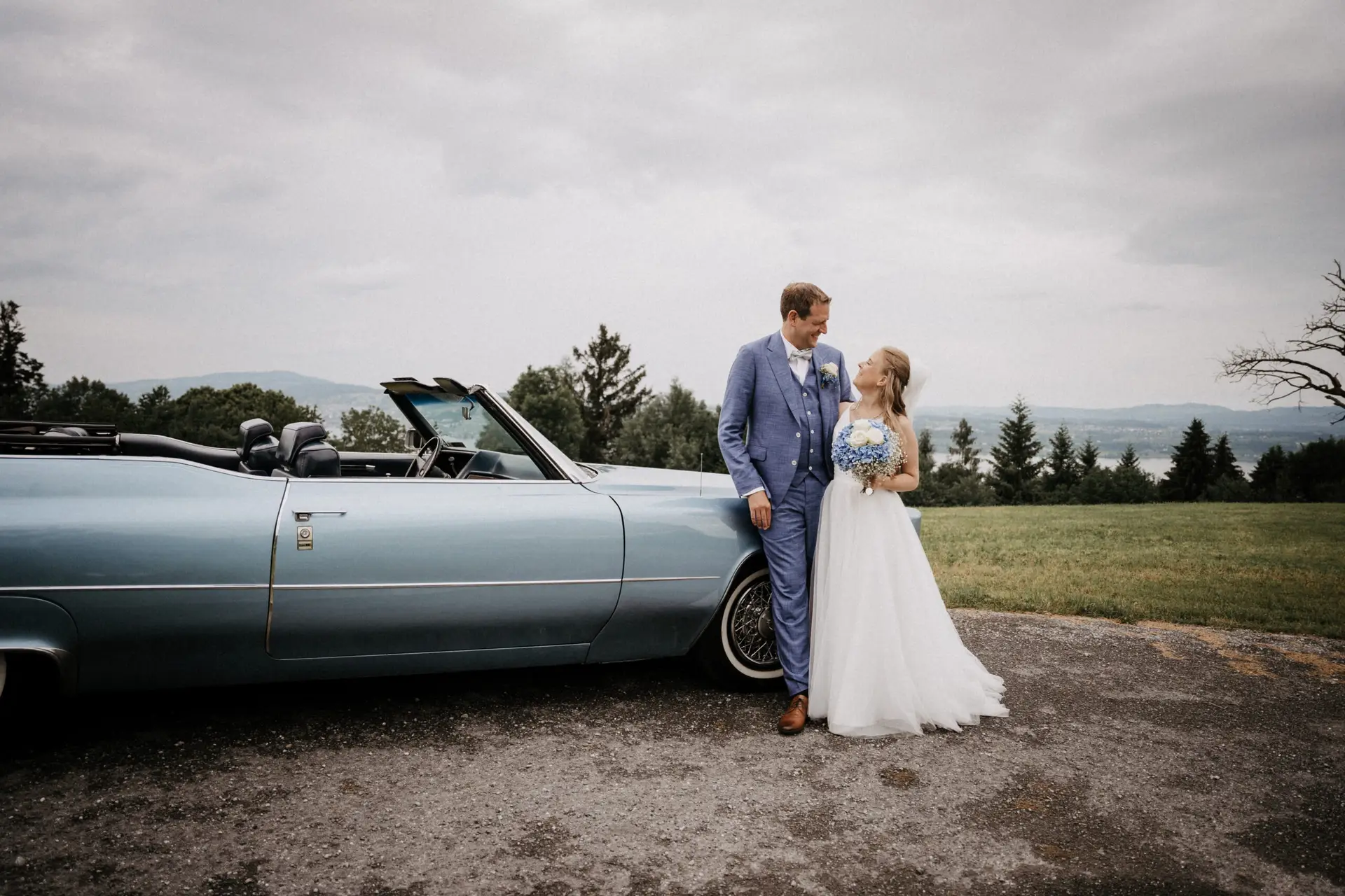 Hochzeitsfoto, Braut und Bräutigam sehen sich verliebt in die Augen und lehnen an wunderschön hellblauen Cabrio vom malerische Kulisse